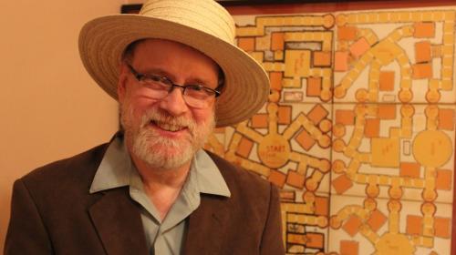 David R. Megarry smiling in a suit coat and straw hat in front of his original Dungeon! board