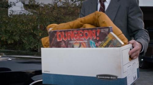 A screenshot from the TV show "Stranger Things," showing a person carrying a box of items including a copy of the board game "Dungeon!"
