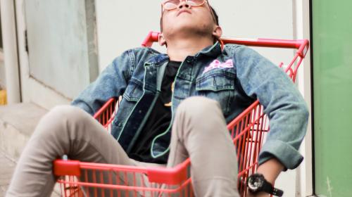 An exhausted adult man lying in a shopping cart, with his head tilted back and limbs draped over the sides.