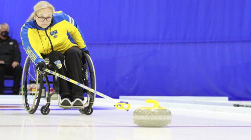 Photo of a woman in a wheelchair playing a game of curling. She’s using a delivery stick to slide, or “throw,” a granite stone into the field of play on the ice.