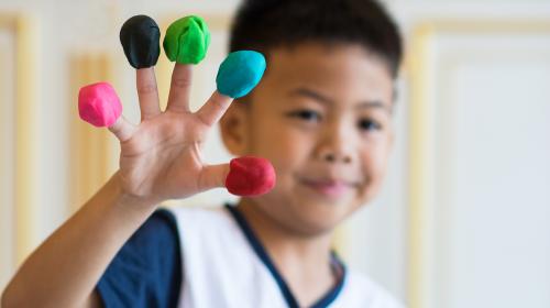 A child holds up one hand, showing a clump of playdoh on the tip of each finger. Each clump is a different color.