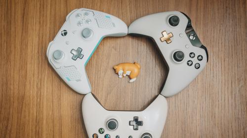 Three XBOX controllers arranged in a circle around a small plastic toy of a shiba inu.