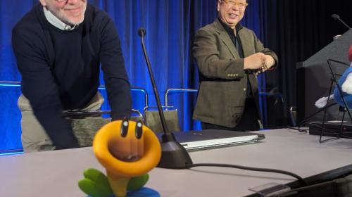 A photo of Takashi Tezuka behind a lectrun, with a Talking Flower figurine on a table in the foregound.