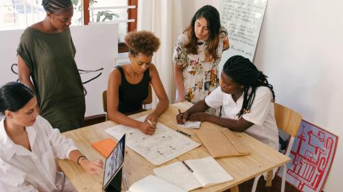 A group of five racially diverse women gather around a table, collaborating on a project.