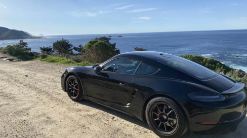 A beautiful photo of Kris's sports car. It is sleek and black on a sandy dirt road with the Pacific ocean and a clear blue sky behind it. Apologies, the writer of this alt text does not know what model the car is.