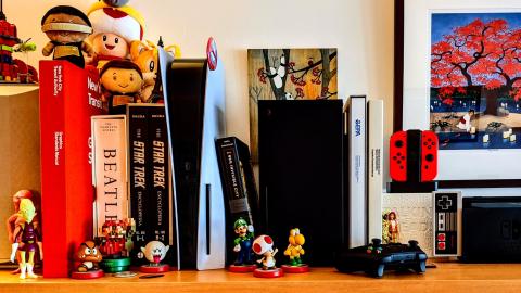 A photo of a PS5 and an Xbox Series X both placed vertically on Mark and Dale's bookshelf, nestled strategically between a series of large reference books.