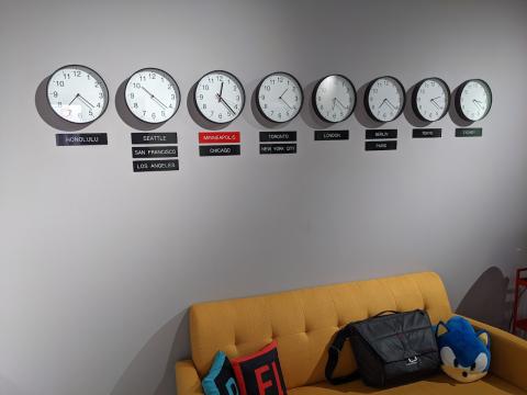 A photo of 8 analog clocks in a row on a gray wall, each depicting a different time zone. Underneath each clock are 1-3 name plaques with the names of world cities.