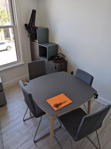 A photo of a square-ish table and chairs in a small conference room. A set of cabinets are in the background, and hold three microphone arms and other audio equipment.
