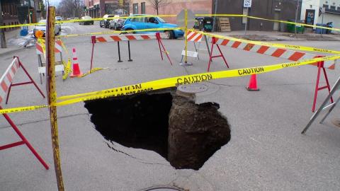 A deep sinkhole in the middle of a city street. The sinkhole has been circled by safety cones and caution tape.