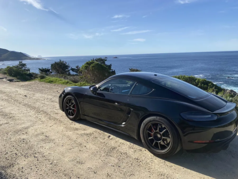 A beautiful photo of Kris's sports car. It is sleek and black on a sandy dirt road with the Pacific ocean and a clear blue sky behind it. Apologies, the writer of this alt text does not know what model the car is.