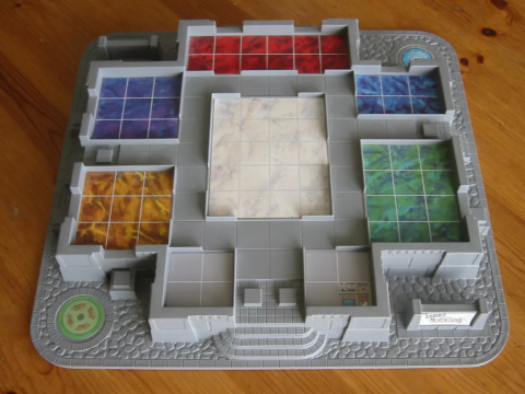 Clue: The Great Museum Caper board. A game board with a grey foundation of a museum build up from it. There are six colored rooms: one in the center, one in the back, and two on each side.