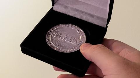 A close-up photo of a large coin in a commemerative box. The coin is silver and features the classic "SEGA" logotype.